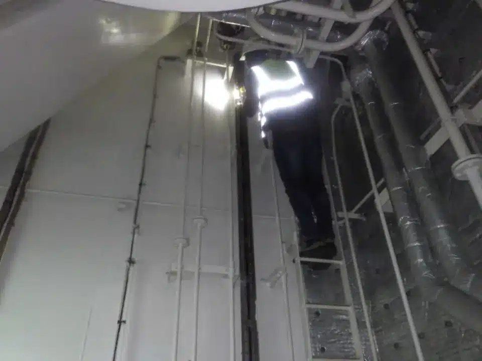 Un technicien portant un gilet de haute visibilité monte une échelle pour une intervention technique dans un espace confiné avec des tuyaux et câblages.